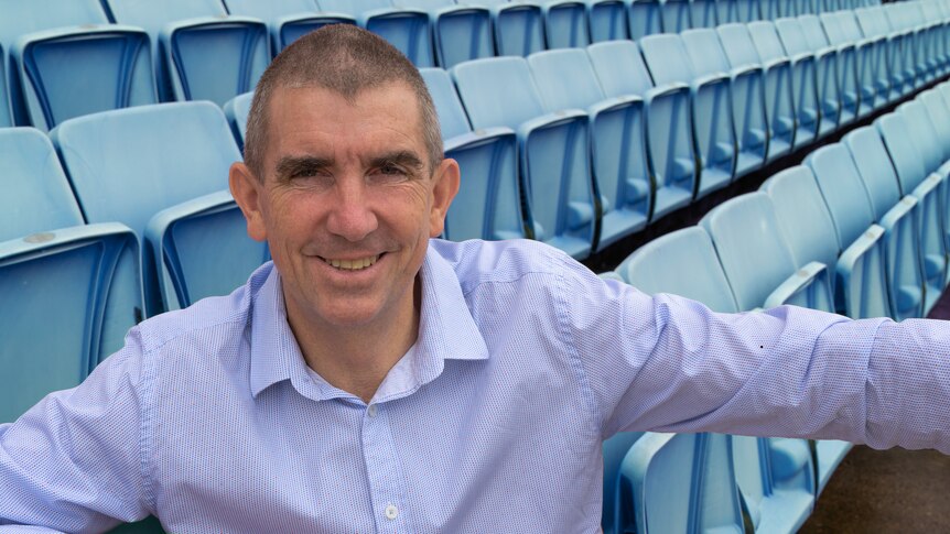 A man with short hair, wearing a lilac shirt, sitting in a grandstand with rows of blue seats in the background.