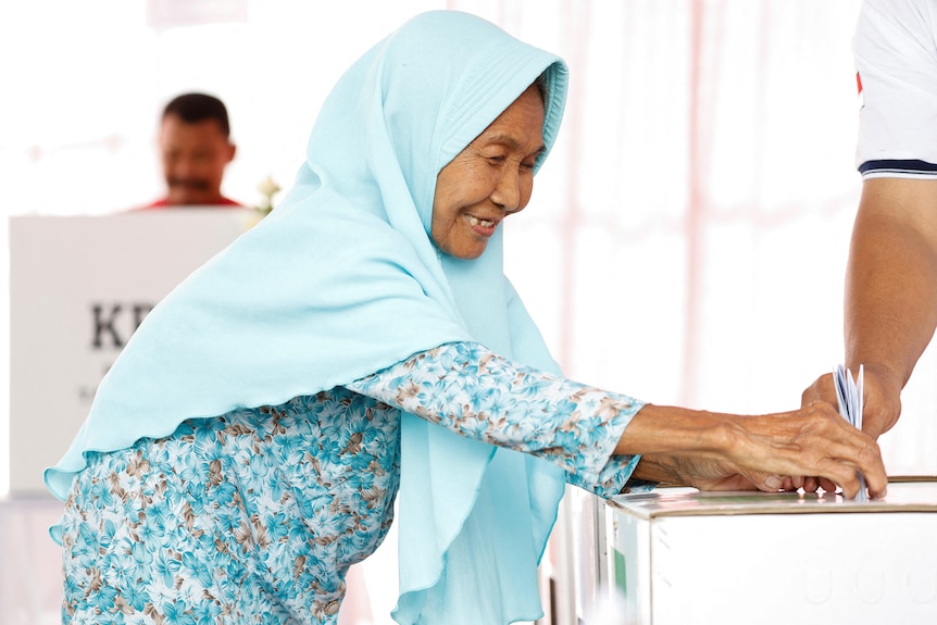 A woman wearing a hijab smiles as she places her ballot in a box.
