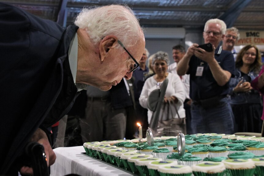 A man stoops down to blow out candles on a cake that says '100' on it. 