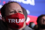 A woman with a red mask bearing the word Trump looks beyond the camera 