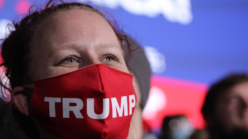 A woman with a red mask bearing the word Trump looks beyond the camera 