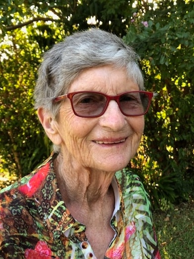 A 87 year old woman in a floral shirt smiles at the camera.