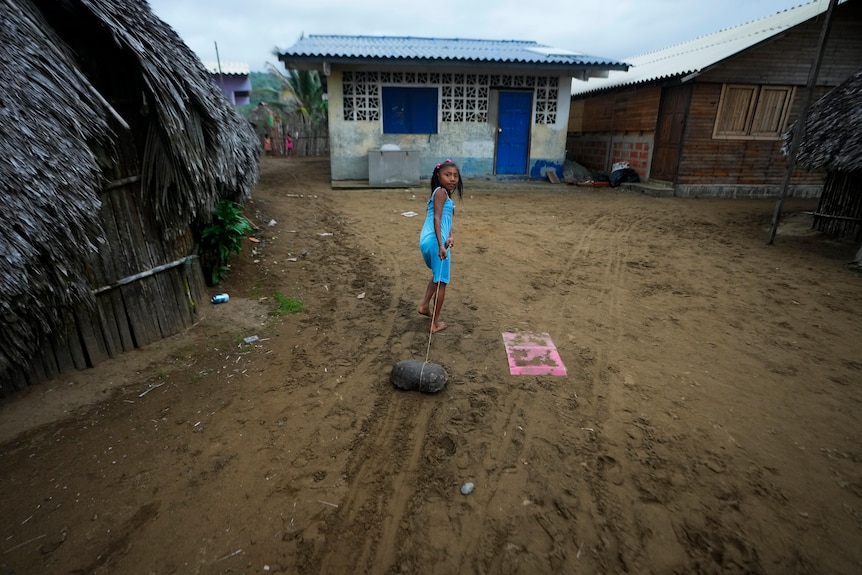 A girl in a blue dress drags a turtle shellwith a rope toward houses in Panama