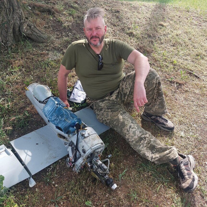 A man in a khaki Tshirt and camouflage reclines on a piece of military equipment