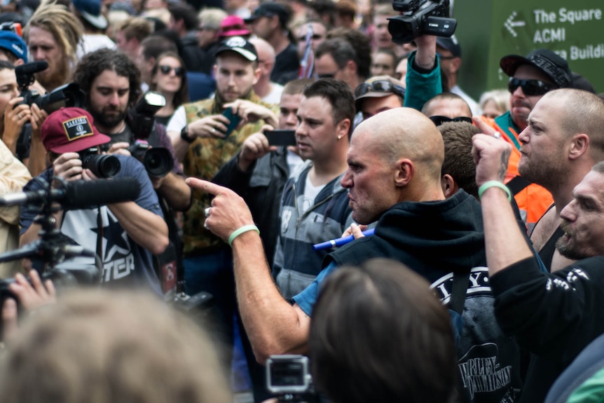 A man with a shaved head points agressivly at a crowd of cameras