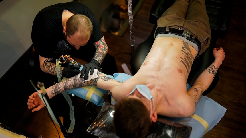 A man in a mask lies on his back getting his arm tattooed. 