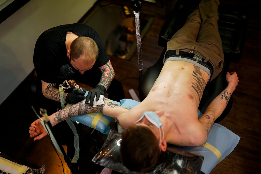 A man in a mask lies on his back getting his arm tattooed. 