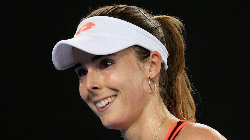 Alize Cornet, wearing an athletic outfit and visor, smiles as she walks on a tennis court.