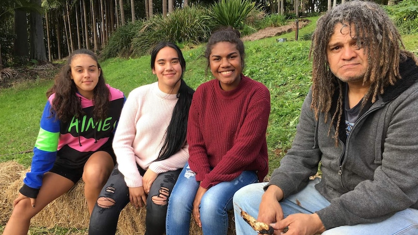 A group of four people sitting on a log in a field with on man with dreadlocks holding a piece of cooked damper.