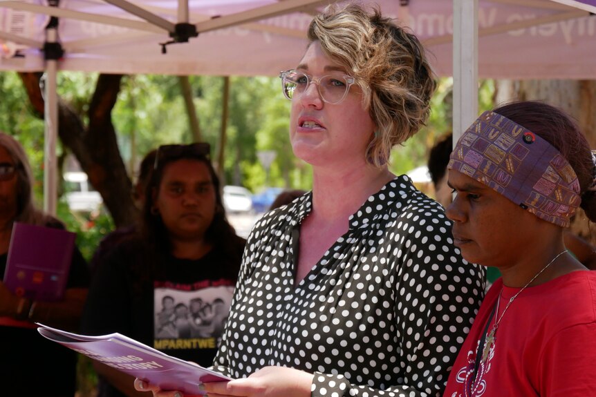 woman with blonde hair wearing polka dots (left) delivering a speech next to woman wearing purple headband and red t-shirt