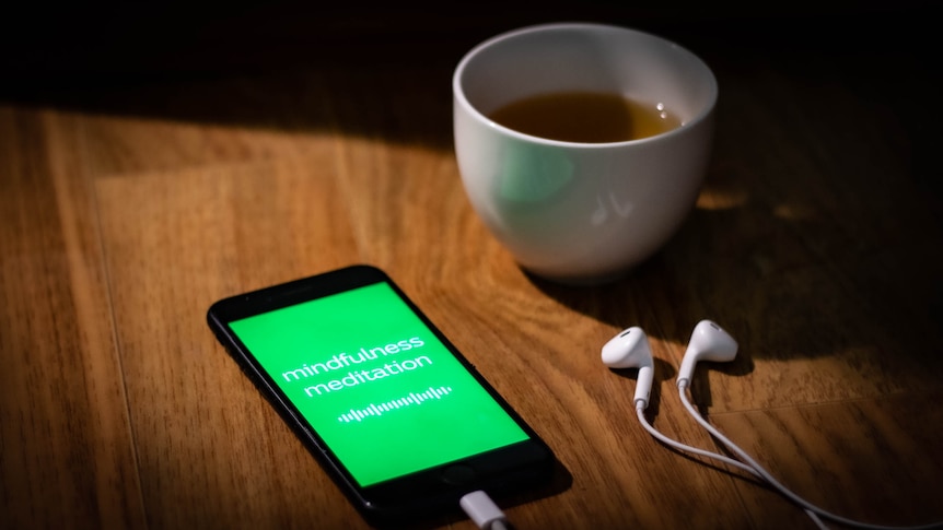 iPhone sits on wood floor next to earbuds. Screen glows green screen with waveform display and words 'mindfulness meditation'.