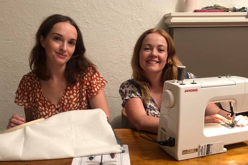 Young brunette woman with a cross-stitch sits next to a young strawberry blonde woman, who is using a sewing machine