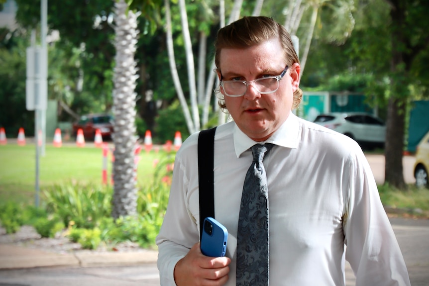 Man with slicked back hear, glasses and a shirt and tie walks with phone in hand and bag over shoulder