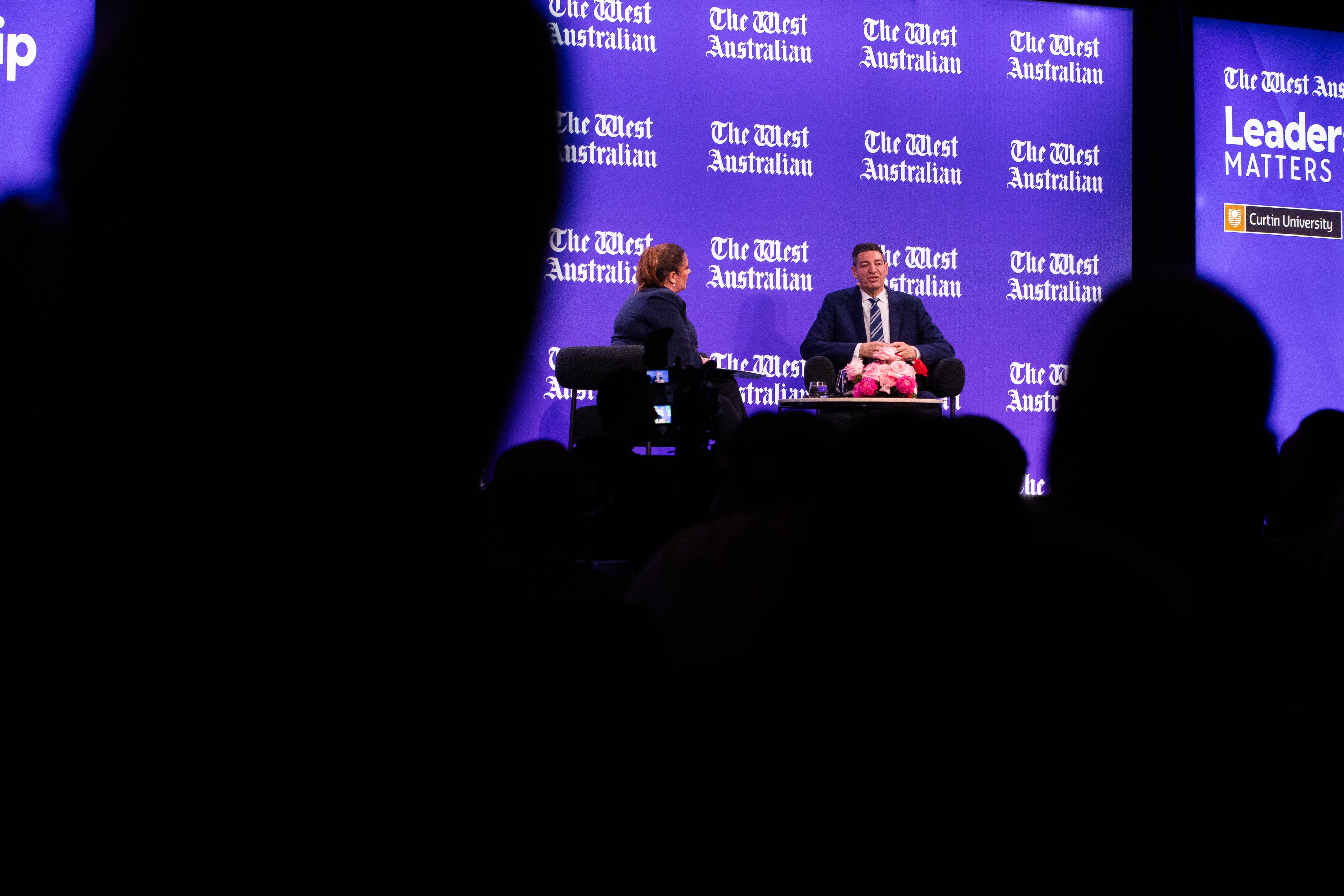 A man sits on stage with a woman, with shadowed audience members in the foreground.