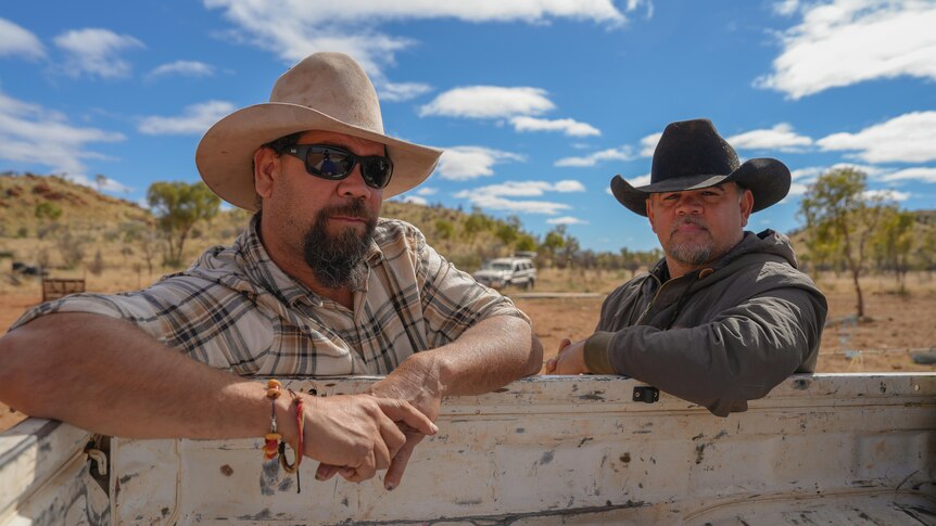 Two stockmen wearing hats lean on the back of a ute and stare into the camera.