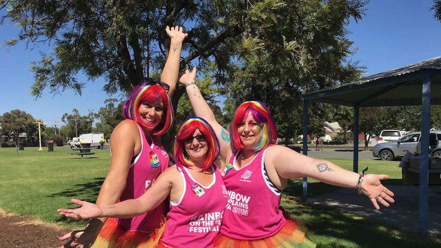 Three women in bright pink wear colourful wigs and strike a flamboyant pose.