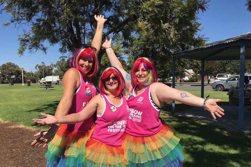 Three women in bright pink wear colourful wigs and strike a flamboyant pose.