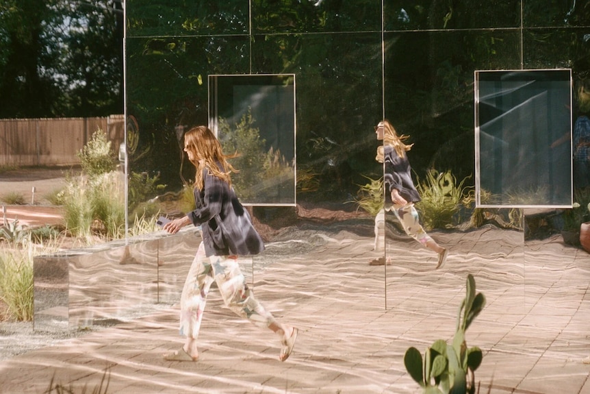 Emma walks in front of a mirrored house with grasses sparsely planted out front and a lone cactus.