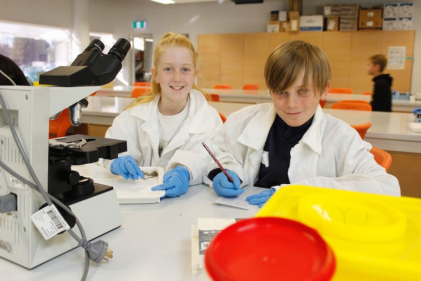 A young boy and girl wearing lab quotes smile at the camera as they work behind a lab table.