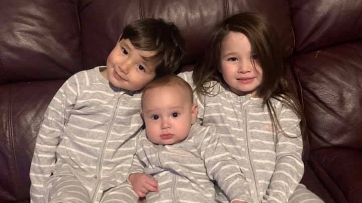 Three young children sit together on a couch and they are all wearing matching grey and white striped onsies.
