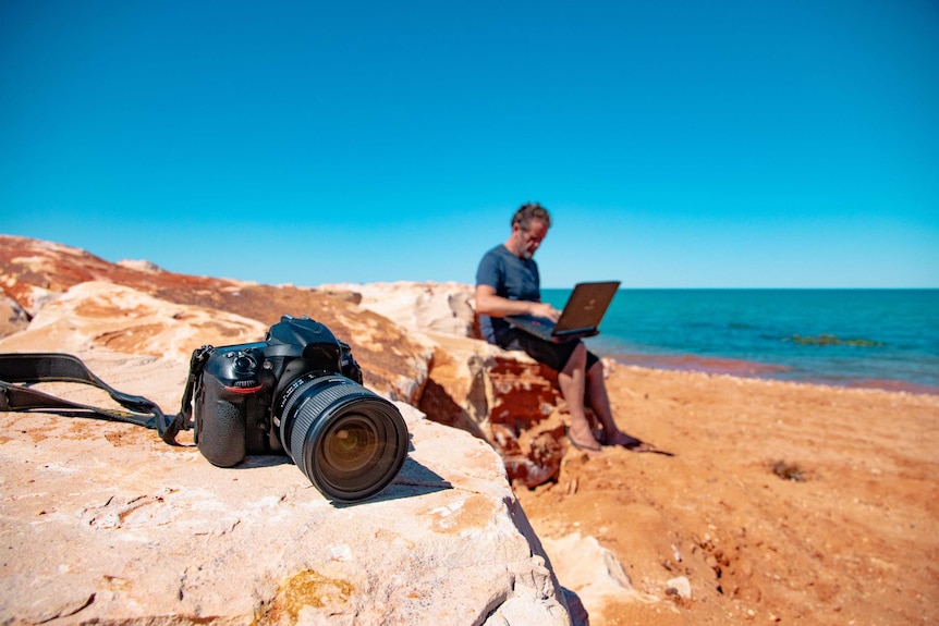 Camera on rock and man behind on laptop, with ocean in background.