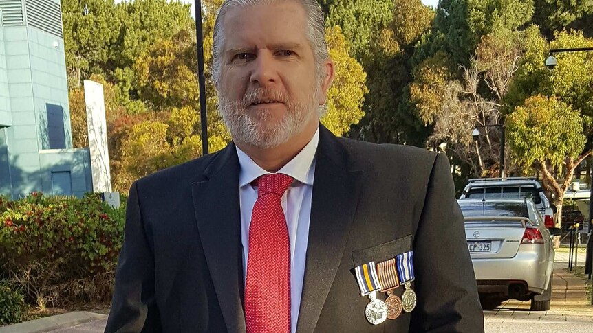A man in a suit with medals pinned on the breast stands in a car park.
