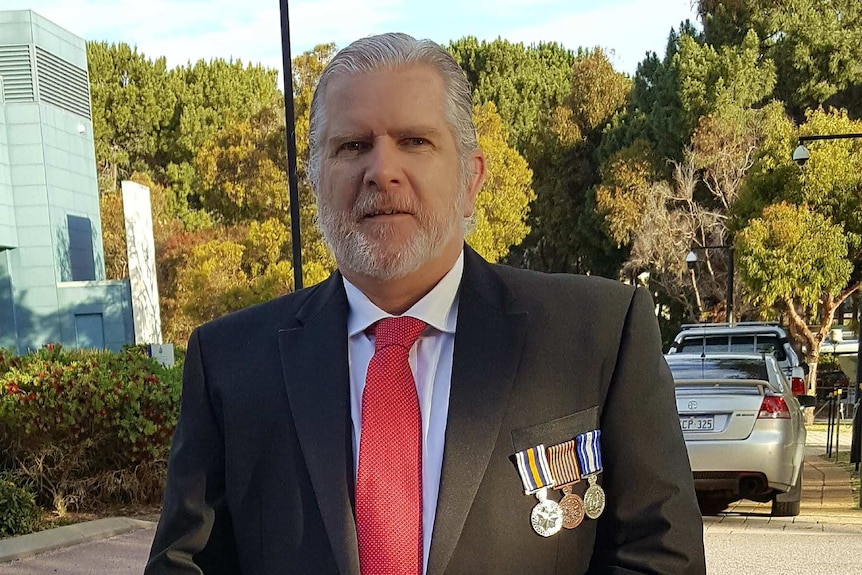 A man in a suit with medals pinned on the breast stands in a car park.