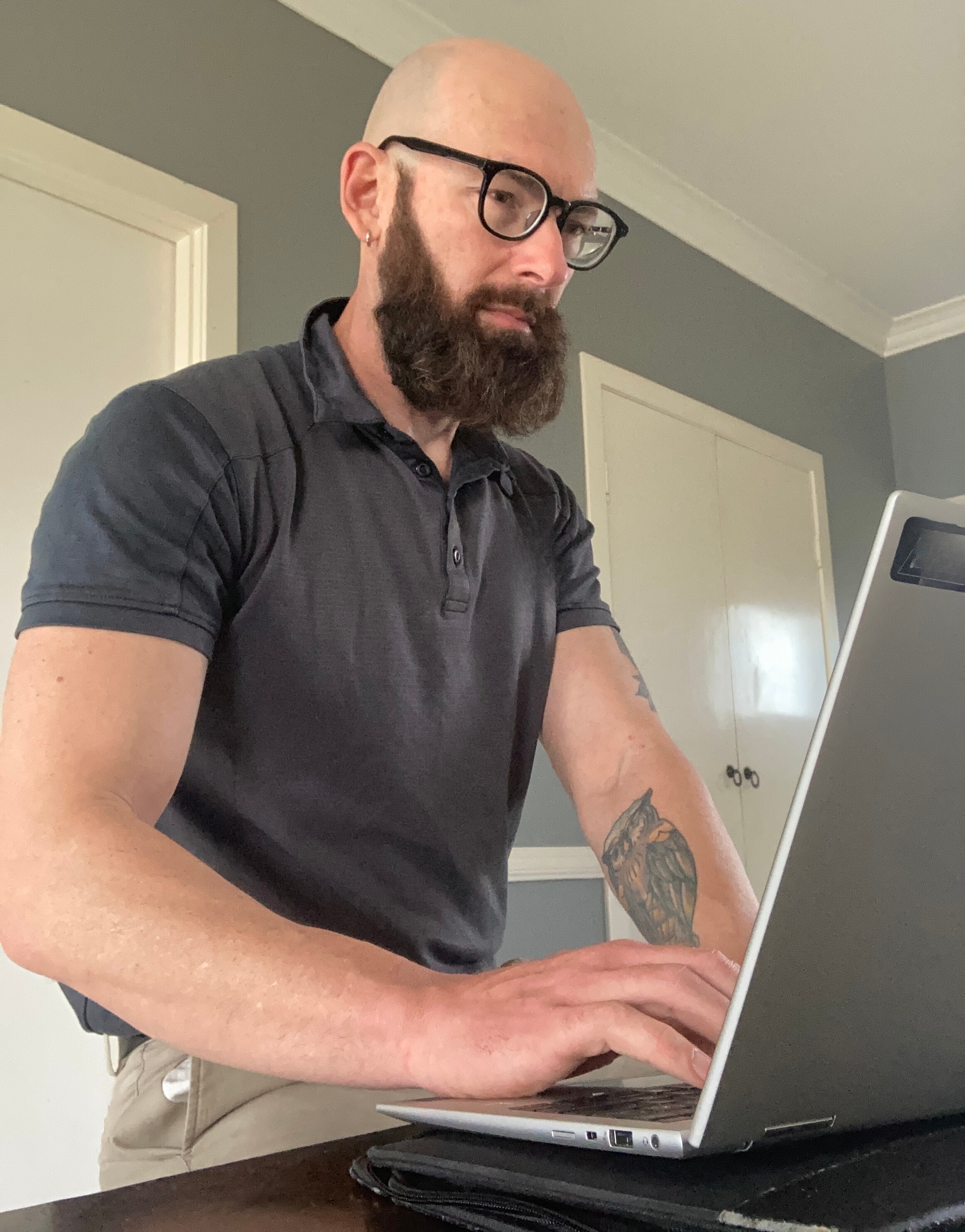 A man in a grey polo with a tattoo on his forearm working on a laptop