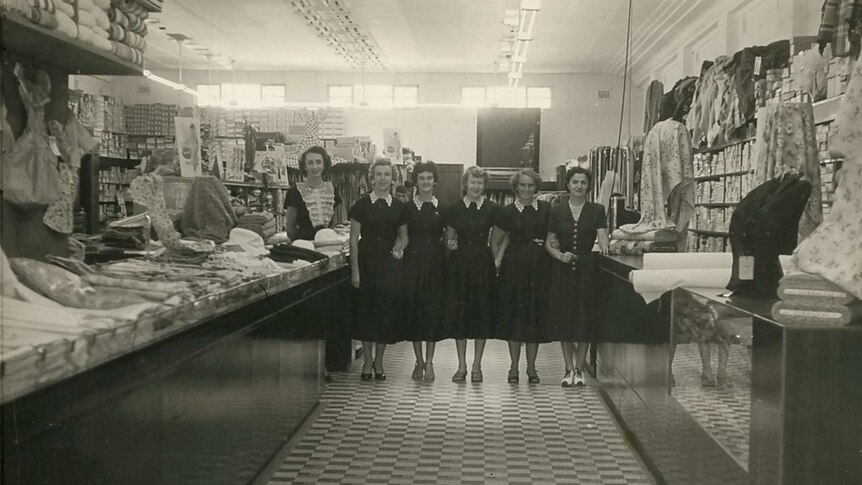 A black and white photo of women standing in an aisle in a ladies department store in 1955.