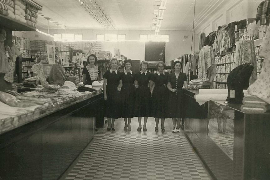 A black and white photo of women standing in an aisle in a ladies department store in 1955.