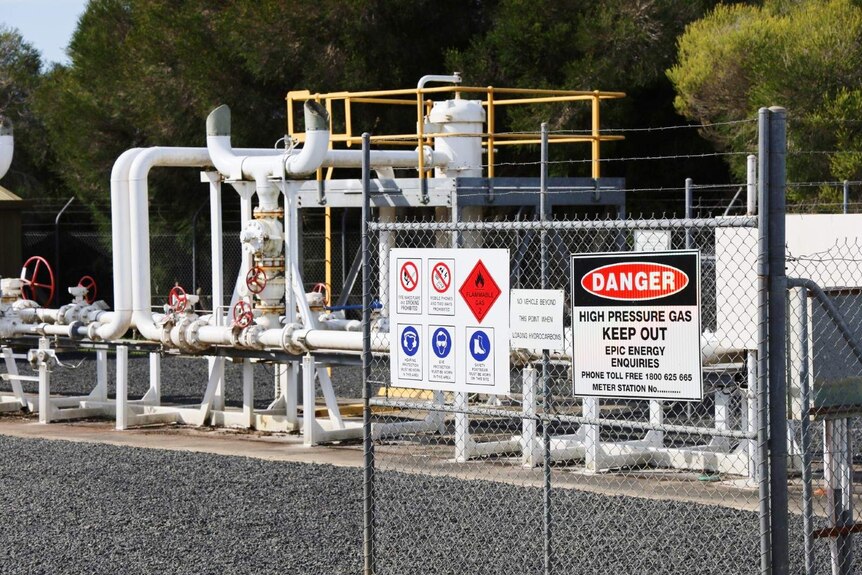 Long white pipe system behind a barbed-wire fence with a sign reading 'Danger. High pressure gas. Keep out.'