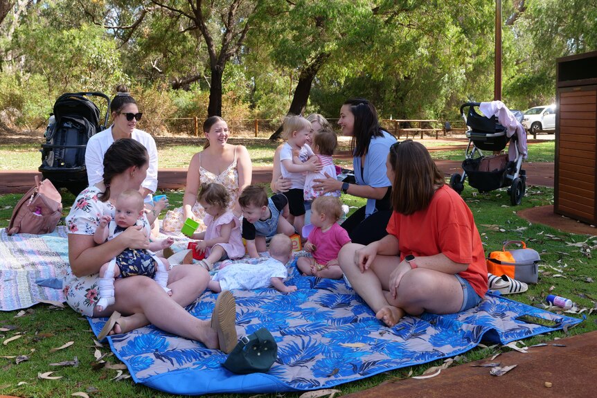 Six mothers sitting together on a picnic rug with seven children sitting and eating.