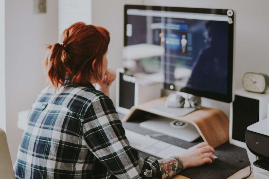 A woman with red hair and a plaid shirt looks at her desktop computer in her home office.