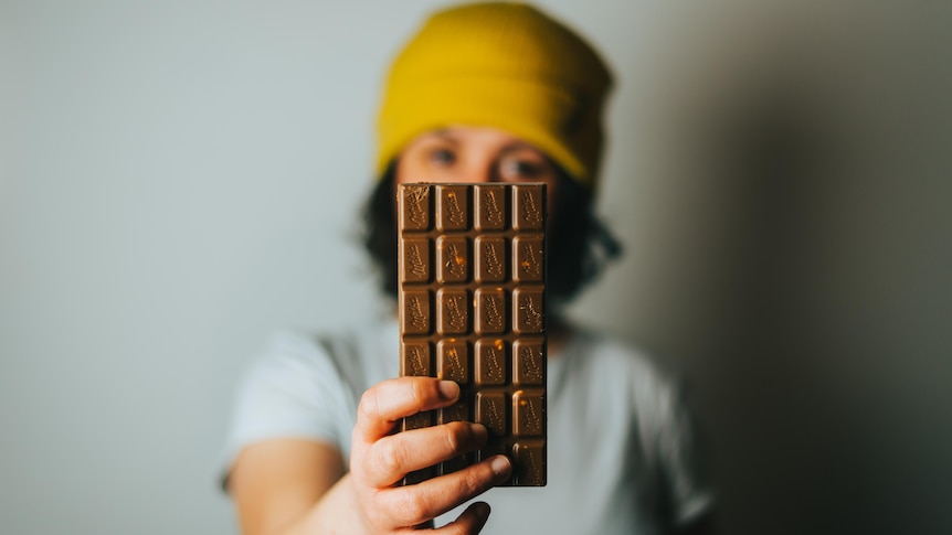 A blurred figure in yellow beanie holds a family-sized chocolate bar out in front of her, taking up the front of field.