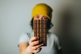 A blurred figure in yellow beanie holds a family-sized chocolate bar out in front of her, taking up the front of field.