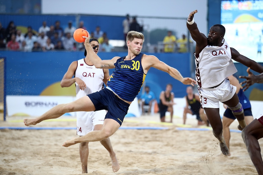 A male beach handball player wears a singlet and shorts as he takes a shot during a match.