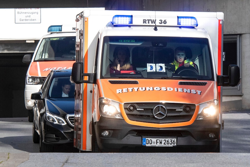 A large Mercedes ambulance in orange colours is followed by a black vehicle and another emergency vehicle.