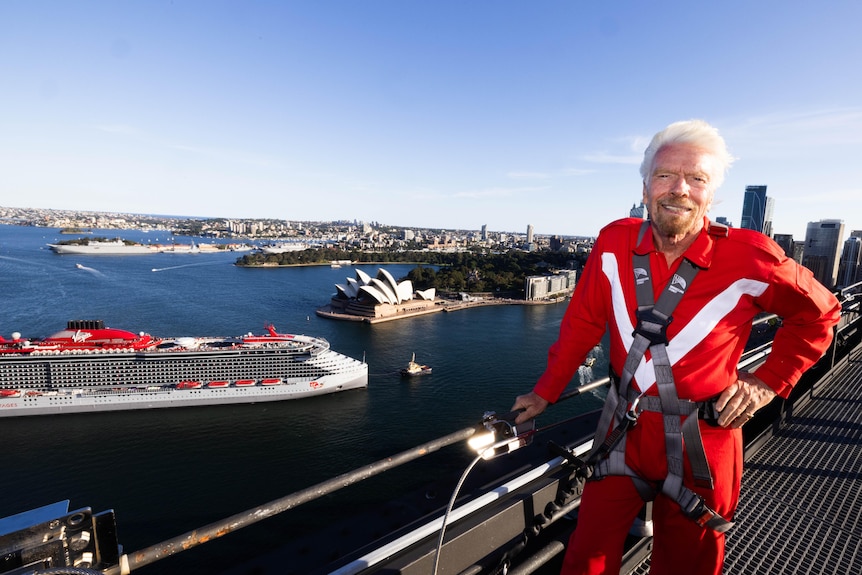 A grey-haired man with a dark beard in a red suit leans on a bridge railing with a cruise ship in the background.