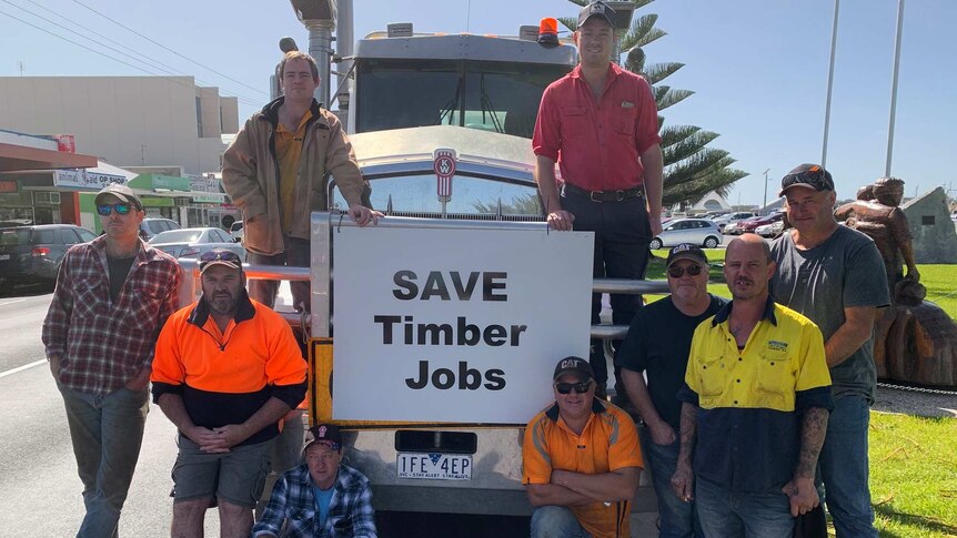 Nine men stand on and next to a transport truck on a sunny day around a sign that says Save Timber Jobs.