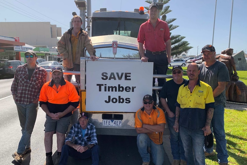 Nine men stand on and next to a transport truck on a sunny day around a sign that says Save Timber Jobs.