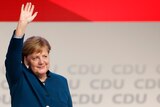 Angela Merkel stands on a stage in front of CDU signage. She waves to the crowd with a smile on her face.