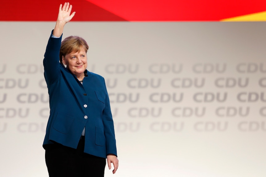 Angela Merkel stands on a stage in front of CDU signage. She waves to the crowd with a smile on her face.