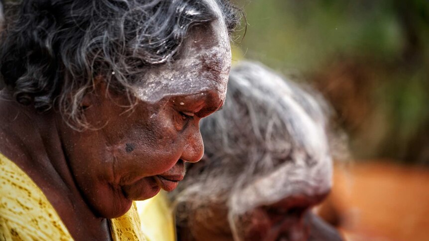 An Aboriginal woman dressed in yellow and wearing white pigment on her forehead looks down.