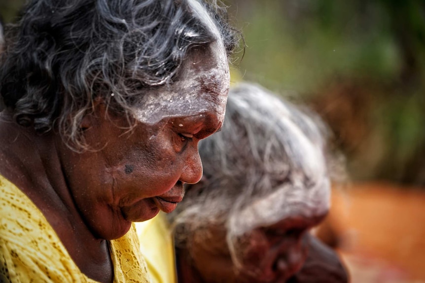 An Aboriginal woman dressed in yellow and wearing white pigment on her forehead looks down.