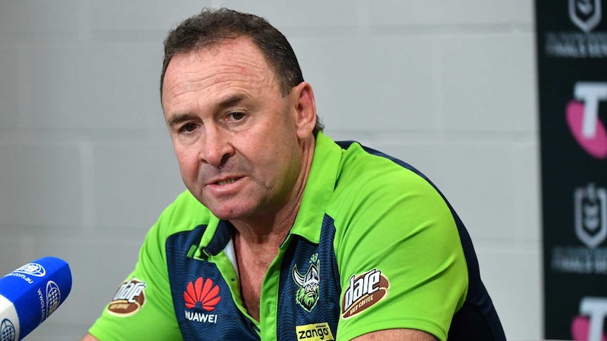 Glum-looking NRL coach dressed in green stares down as he answers a question at a press conference.