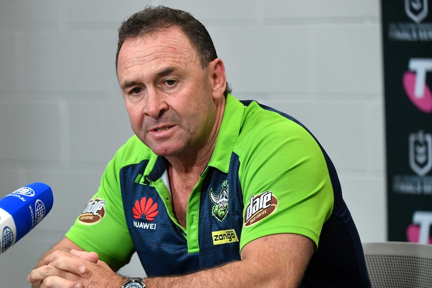 Glum-looking NRL coach dressed in green stares down as he answers a question at a press conference.