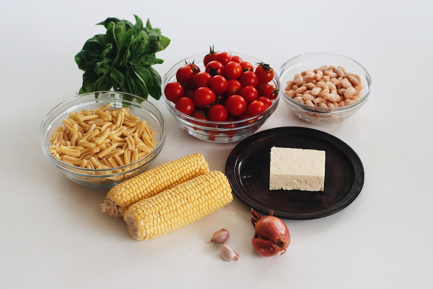 Ingredients for pasta salad on a white bench: basil, tomatoes, a bowl of butter beans, feta, onion, garlic, corn, pasta.