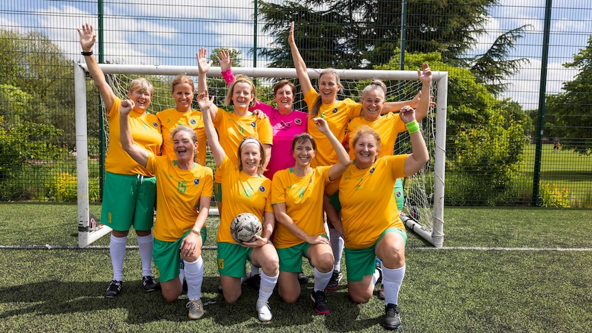 A group of older women in green and gold soccer get up kneeling in front of a soccer goal