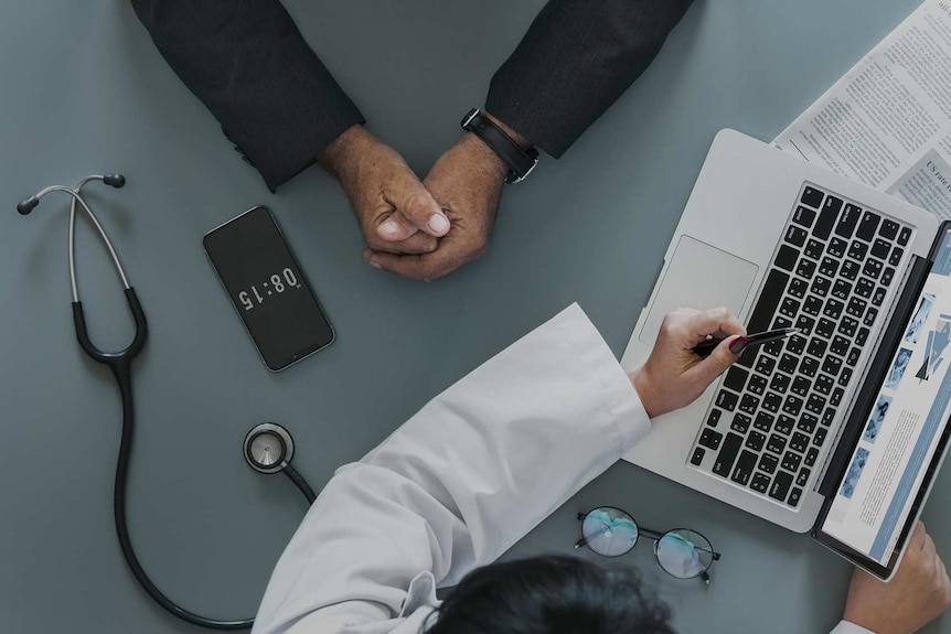 A doctor's hand points at a laptop. A stethoscope sits on the desk. Another pair of hands are clasped opposite the laptop.