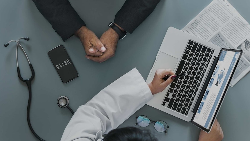 A doctor's hand points at a laptop. A stethoscope sits on the desk. Another pair of hands are clasped opposite the laptop.